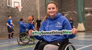 Young woman in sports wheelchair with basketball court in background holds green sign whiich reads 'Try something new'
