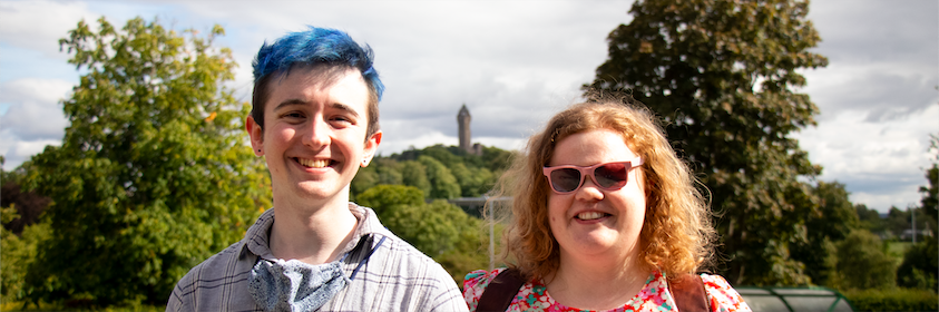 A picture containing smiling young male personal assistant and female visually impaired employer, outdoor, trees and tower Wallace monument on hillside