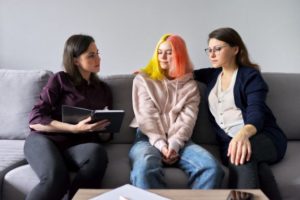 A woman with a clipboard sits on a sofa with a younger woman with pink and yellow hair and an older woman