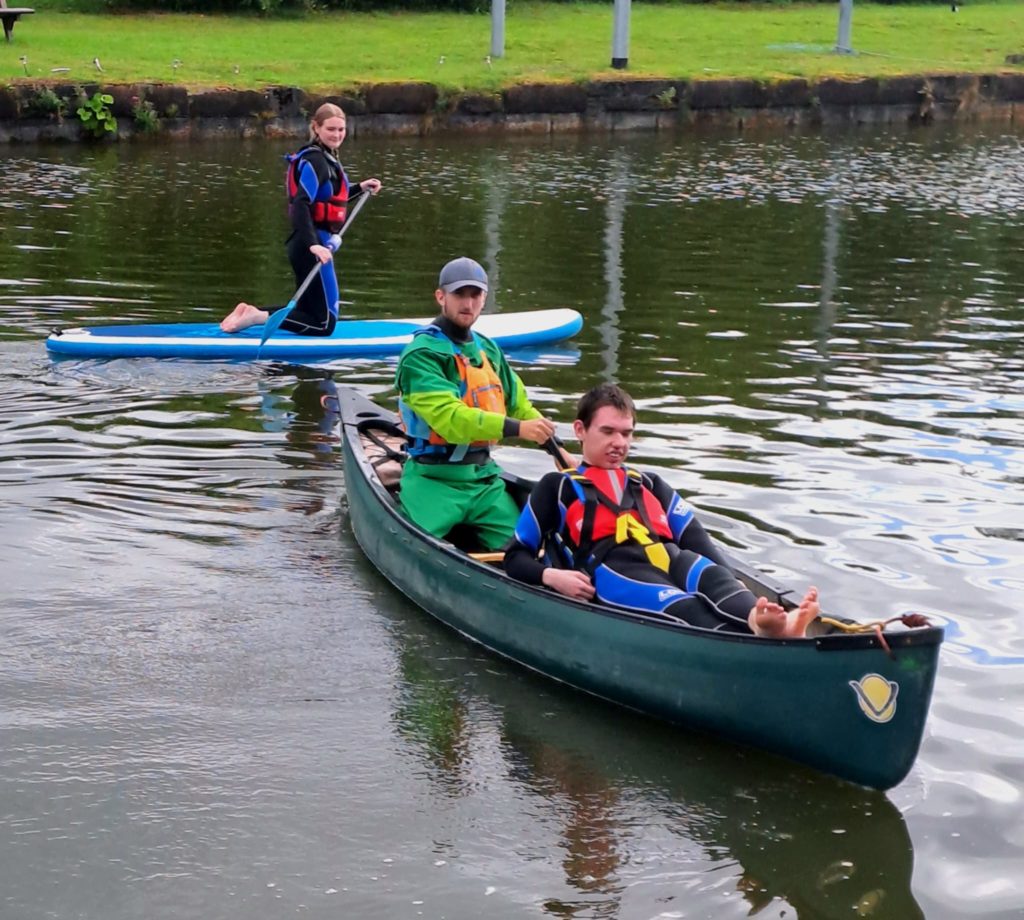 A young man reclines in a canoe, the instructor is sat behind him paddling and a young woman is on a paddleboard behind