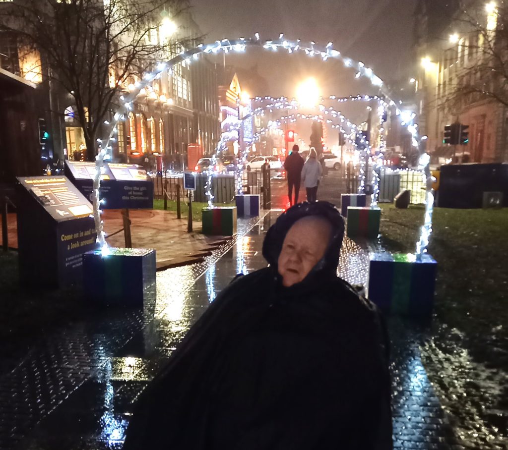 A man wearing a hooded coat in the foreground with Christmas lights on an Edinburgh street behind