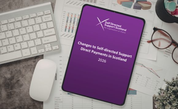 A smart tablet with the front cover of SDSS' "Changes to Self-directed Support Direct Payments in Scotland" report displayed, sitting on top of papers with a keyboard and mouse to the left and glasses to the right, on top of a dark marble surface